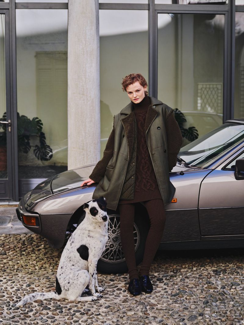 A stylish woman in olive outerwear poses with a black and white dog next to a grey Porsche on a cobblestone driveway near a building.