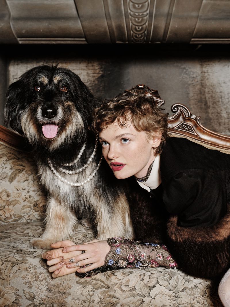 Woman and dog pose on vintage sofa. The woman wears a dark jacket with floral-embellished sleeves. The dog wears a pearl necklace.
