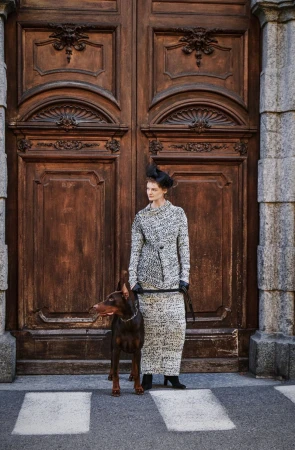 Adult woman in a textured tweed jacket and skirt stands next to a Doberman Pinscher dog in front of large wood doors outdoors.