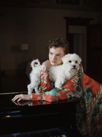 Woman in patterned top poses indoors with two fluffy white dogs near a black piano, creating a cozy and stylish scene.