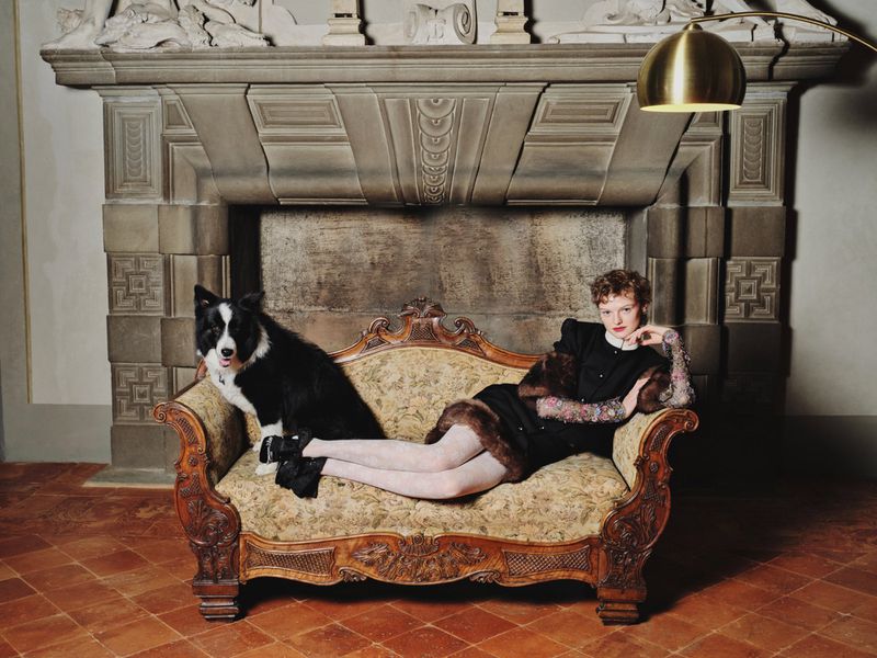 Black and white dog poses with a woman in a black dress on an ornate couch, set against an elaborate fireplace mantel.