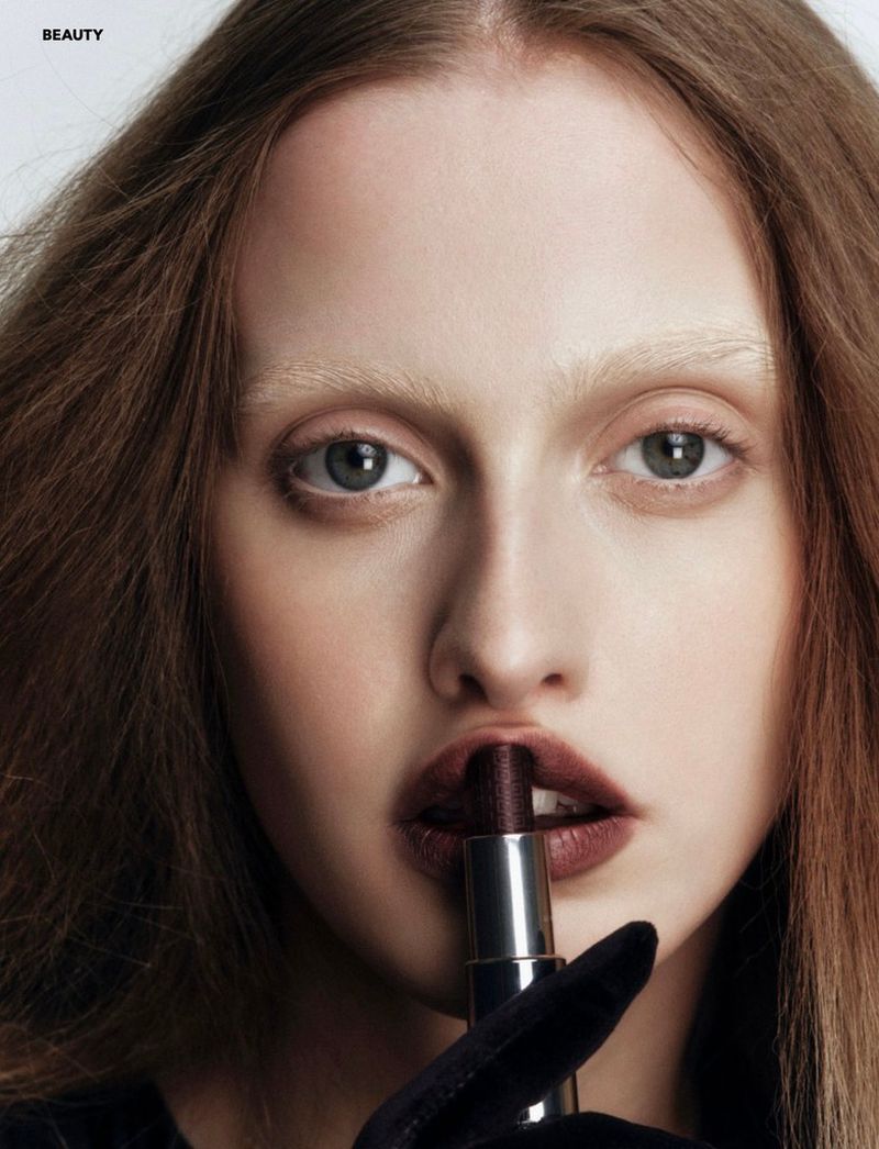 Close-up of a young woman with auburn hair holding a dark lipstick to her lips in a beauty shot against a neutral backdrop.