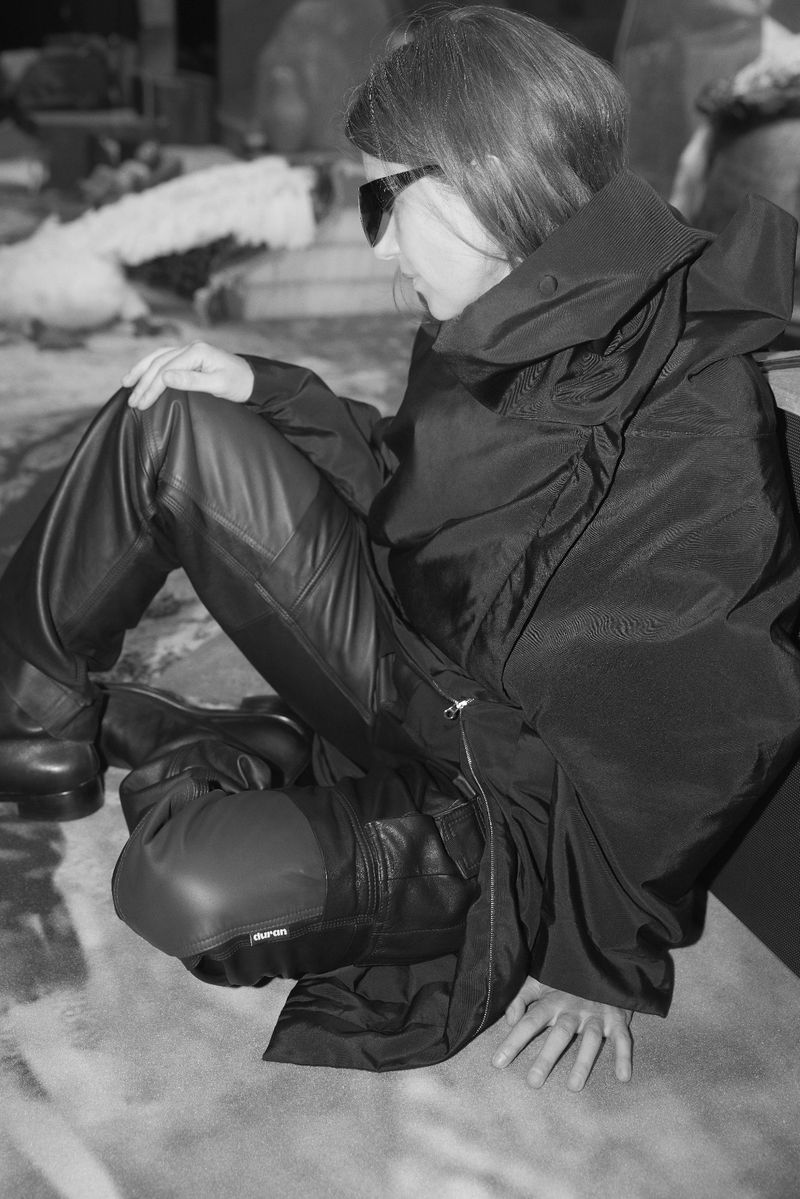 A woman in a dark outfit and sunglasses is seated on a mottled surface, wearing leather pants and a voluminous black coat.