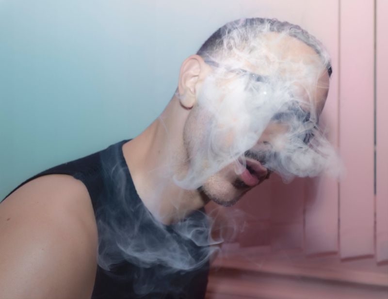 Man with smoke obscuring his face in a close-up shot with a light-blue and pink background, wearing a black tank top.