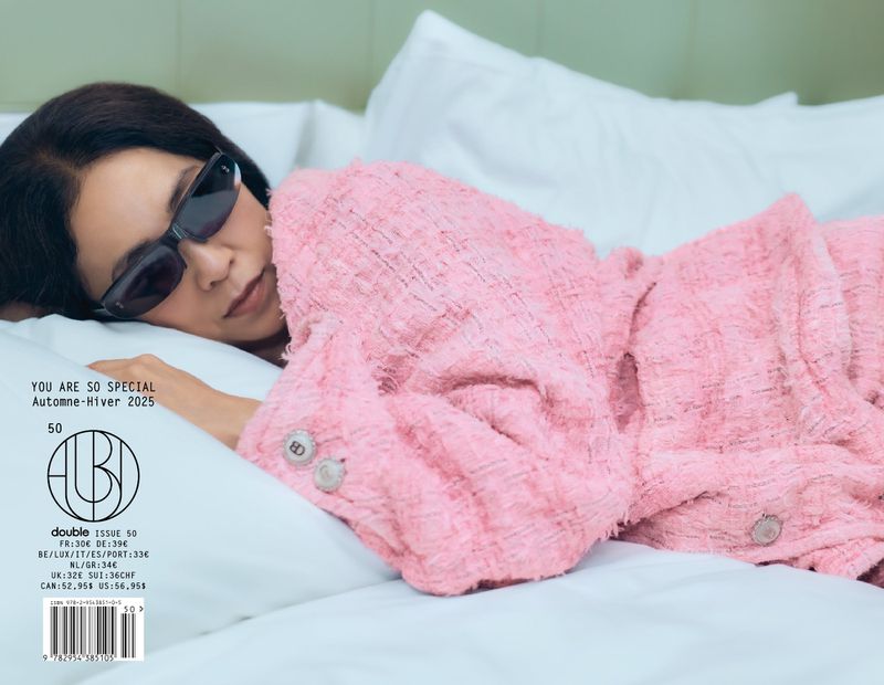 Magazine cover features an adult woman with sunglasses lying on white bedding, wearing a pink tweed jacket. 