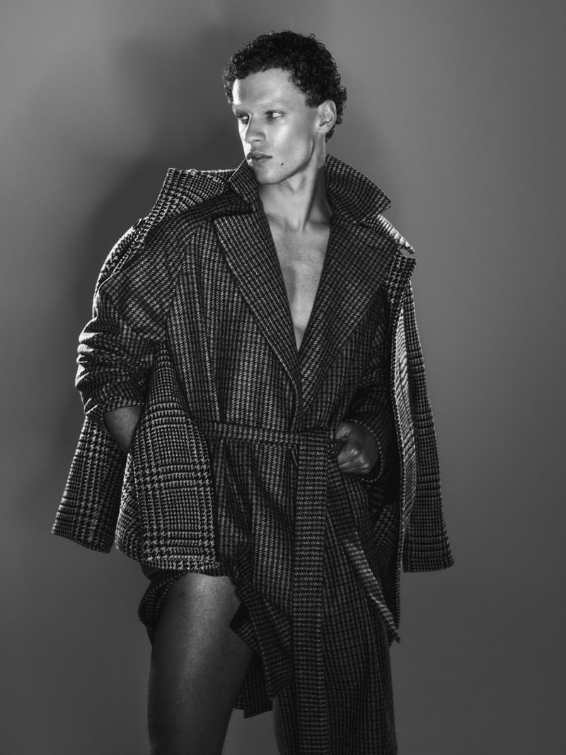 Black and white portrait of a shirtless man wearing a plaid coat looking left in a studio. Fashion shot with dark background.