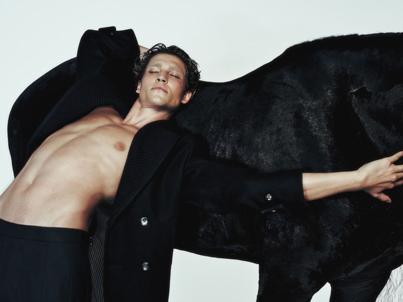 A shirtless man in a coat poses with a black horse against a white backdrop, highlighting fashion and animal themes.