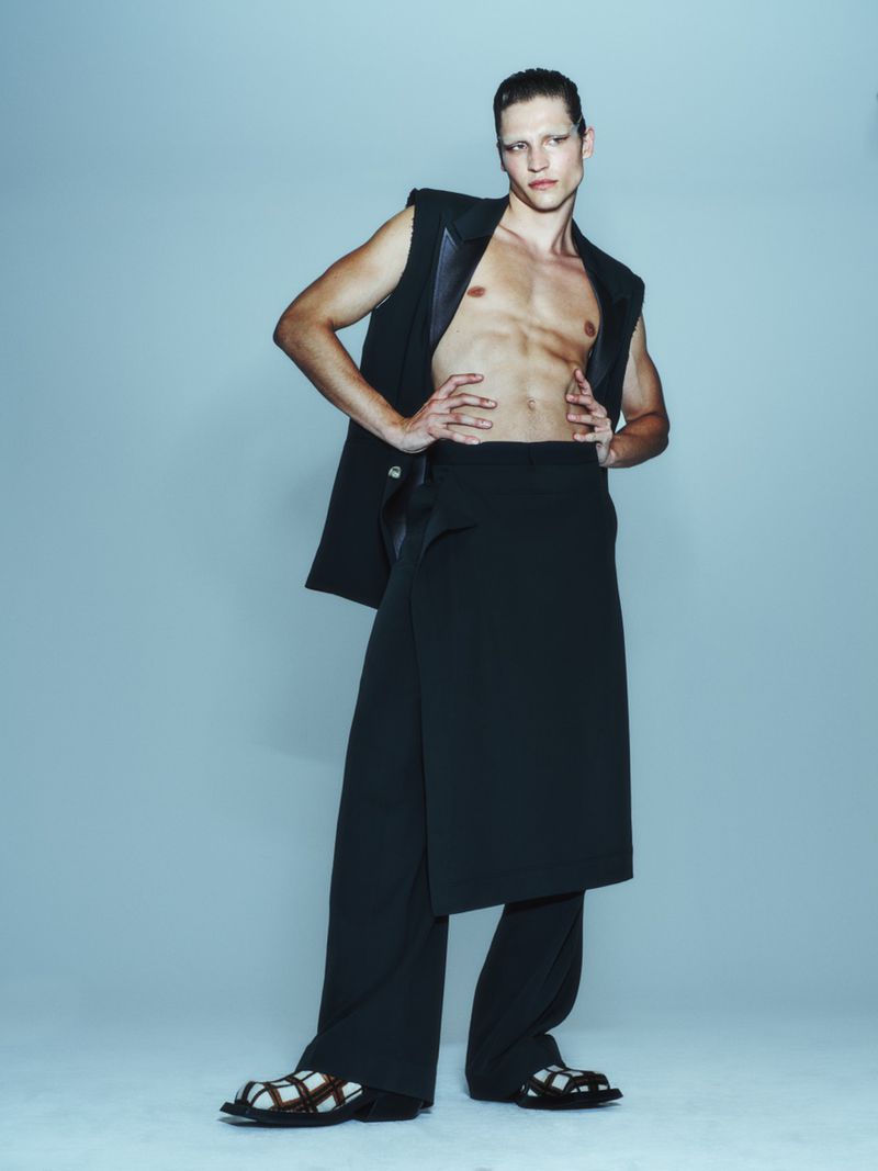 Full-length portrait of a man wearing a sleeveless black jacket, wraparound pants, and sandals in a studio setting, against a light blue background.