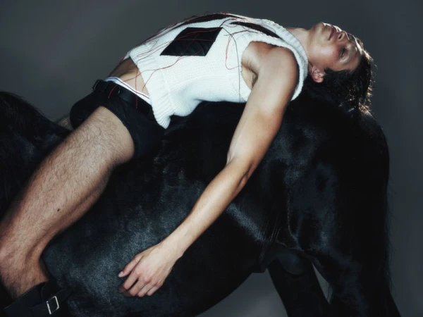 Male fashion model reclines on a black horse in a studio. He wears a white sweater and black shorts, showcasing fashion and equine themes.