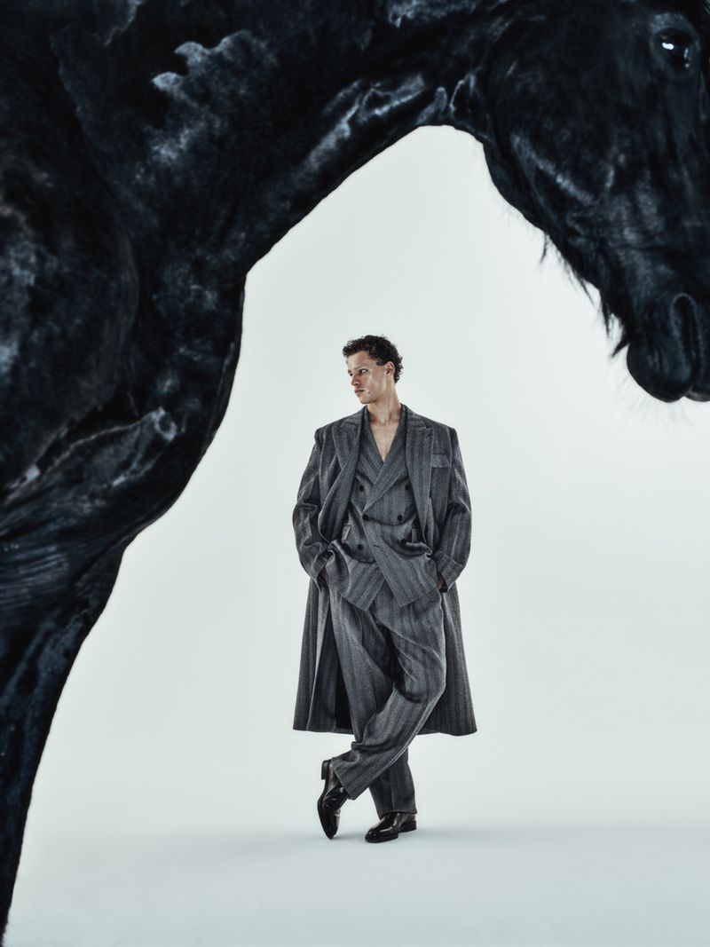 A man poses in a grey pinstripe suit and coat in front of a large black horse prop against a seamless studio backdrop.