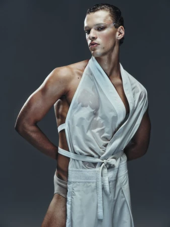 Portrait of a muscular adult male with wet hair and makeup wearing a white apron-like garment, hands behind his back, against a gray backdrop.