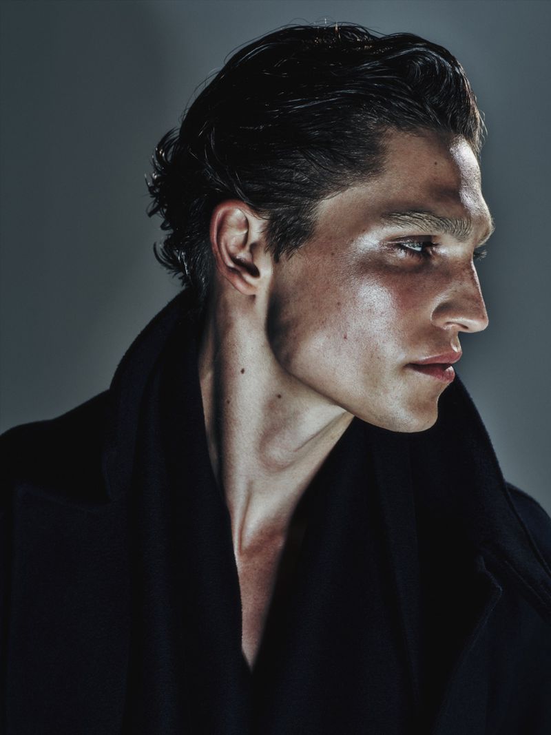 Portrait of a dark-haired man in a black coat looking to the side, with an expressive face highlighted by studio lighting against a gray background.