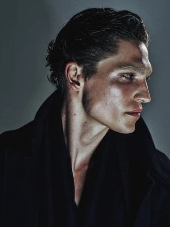 Portrait of a dark-haired man in a black coat looking to the side, with an expressive face highlighted by studio lighting against a gray background.