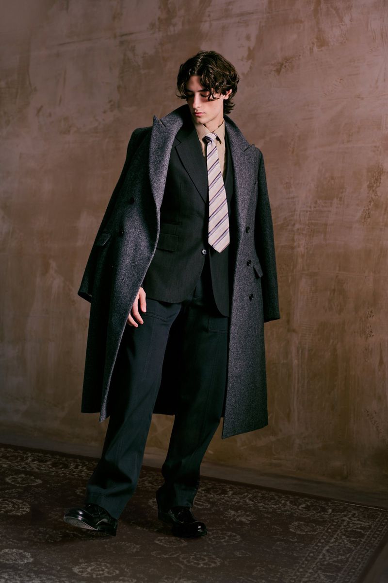 Full-length portrait of a young adult male in a dark suit and grey overcoat, indoors with an aged, textured backdrop.