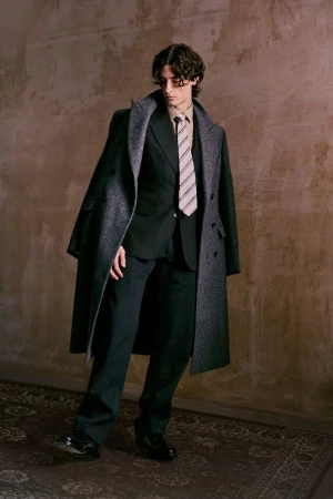 Full-length portrait of a young adult male in a dark suit and grey overcoat, indoors with an aged, textured backdrop.