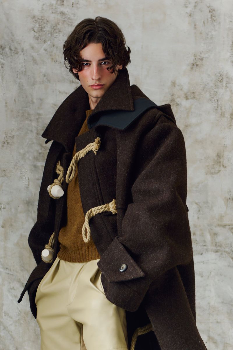 A fair-skinned young adult male with dark hair wearing a brown toggle coat, tan sweater, and tan shorts, posing against a mottled gray background.