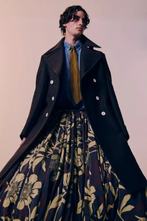 Masculine fashion model wearing a black peacoat, blue dress shirt, patterned gold tie, and a large floral skirt in a studio setting.