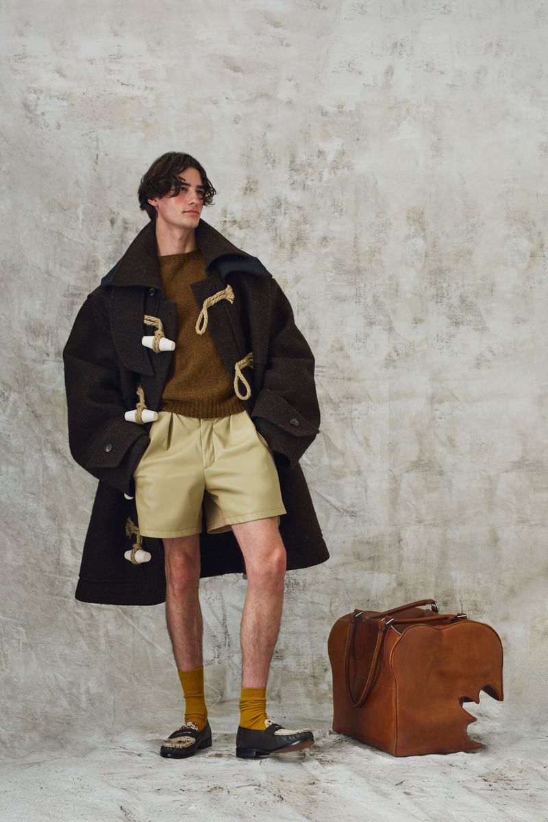 Man posing in an oversized dark duffle coat, pleated beige shorts, and yellow socks next to a brown leather bag on a textured backdrop.