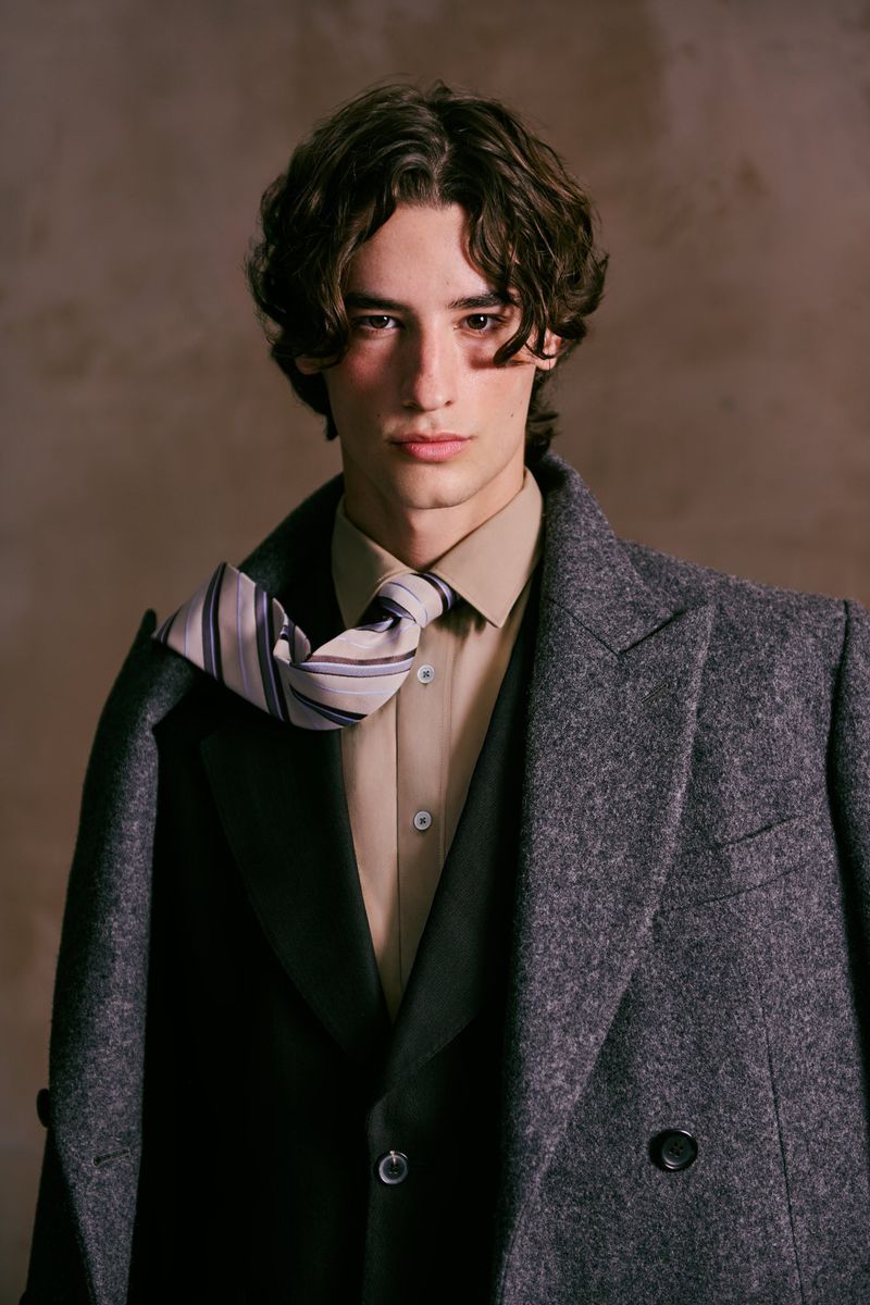 Portrait of a young adult man with dark hair wearing a gray wool coat, beige shirt, and tie, set against a neutral background.