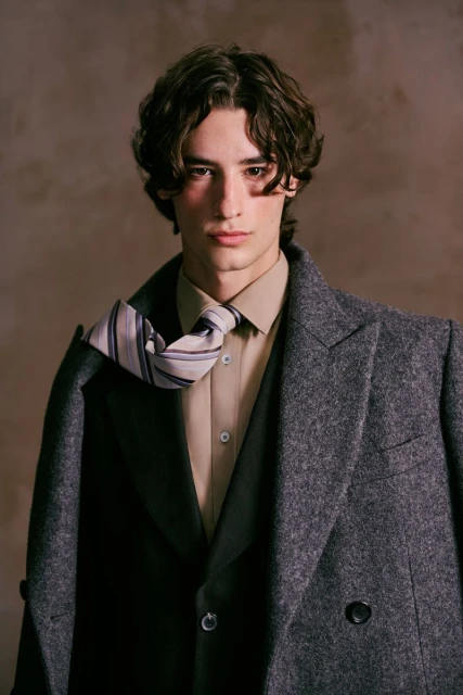 Portrait of a young adult man with dark hair wearing a gray wool coat, beige shirt, and tie, set against a neutral background.