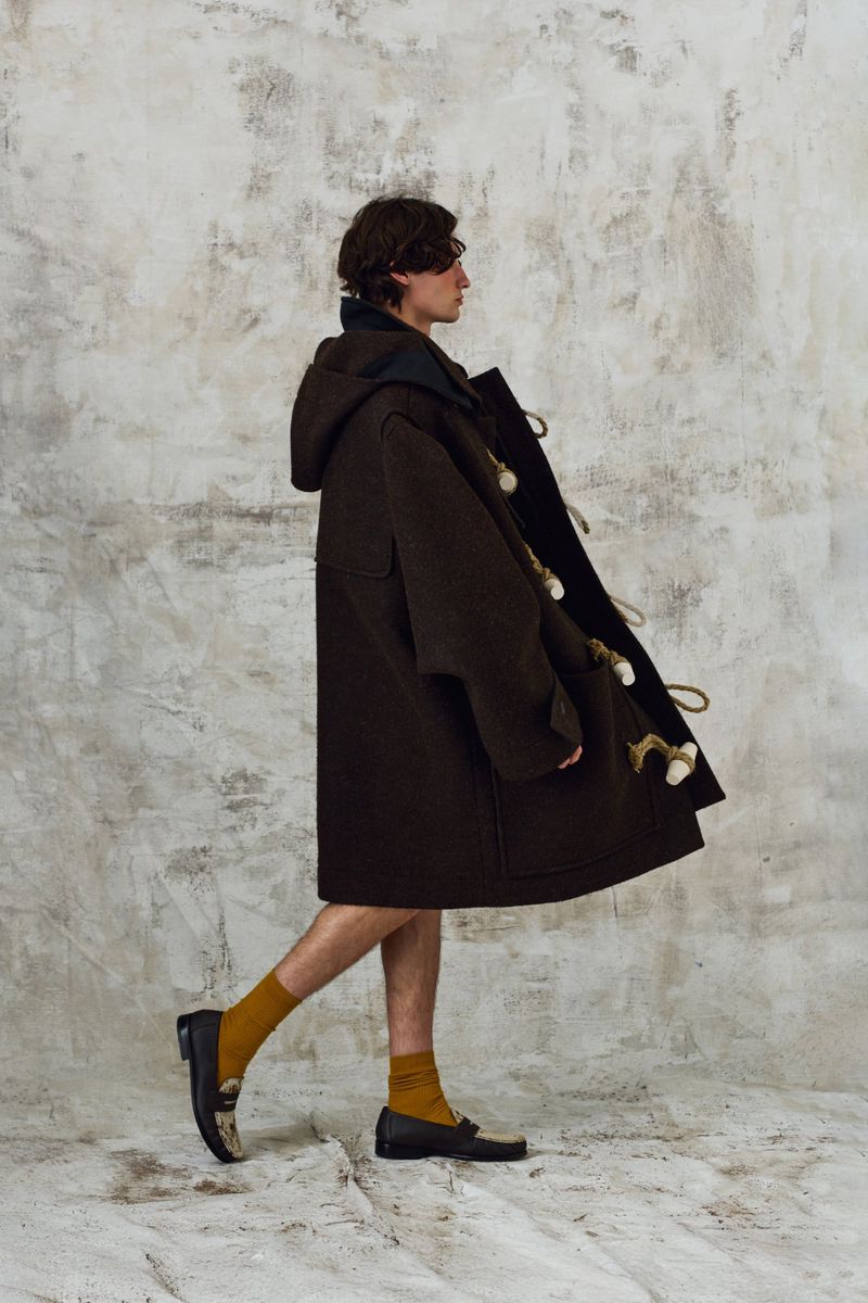 Man in a brown duffle coat walks against a mottled grey backdrop, wearing yellow socks and black loafers in a studio setting.