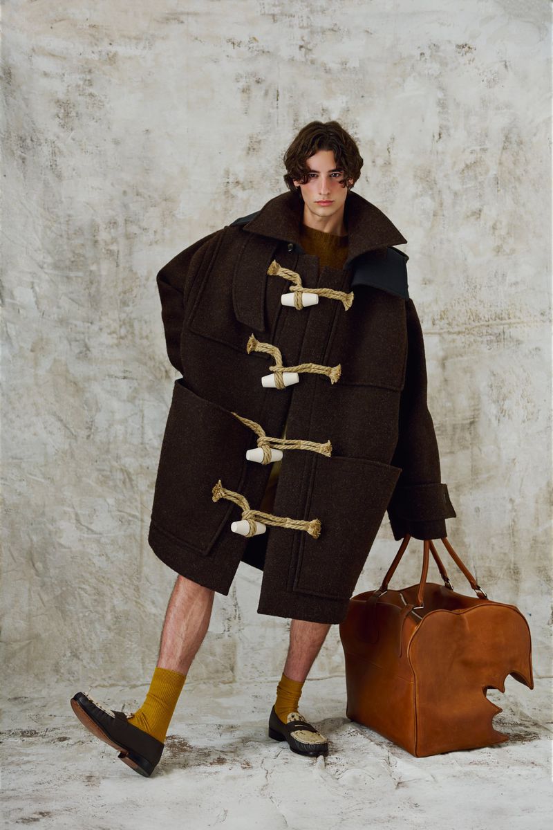 Adult male model poses in an oversized brown toggle coat with tan leather duffel bag in a fashion editorial on a concrete backdrop.
