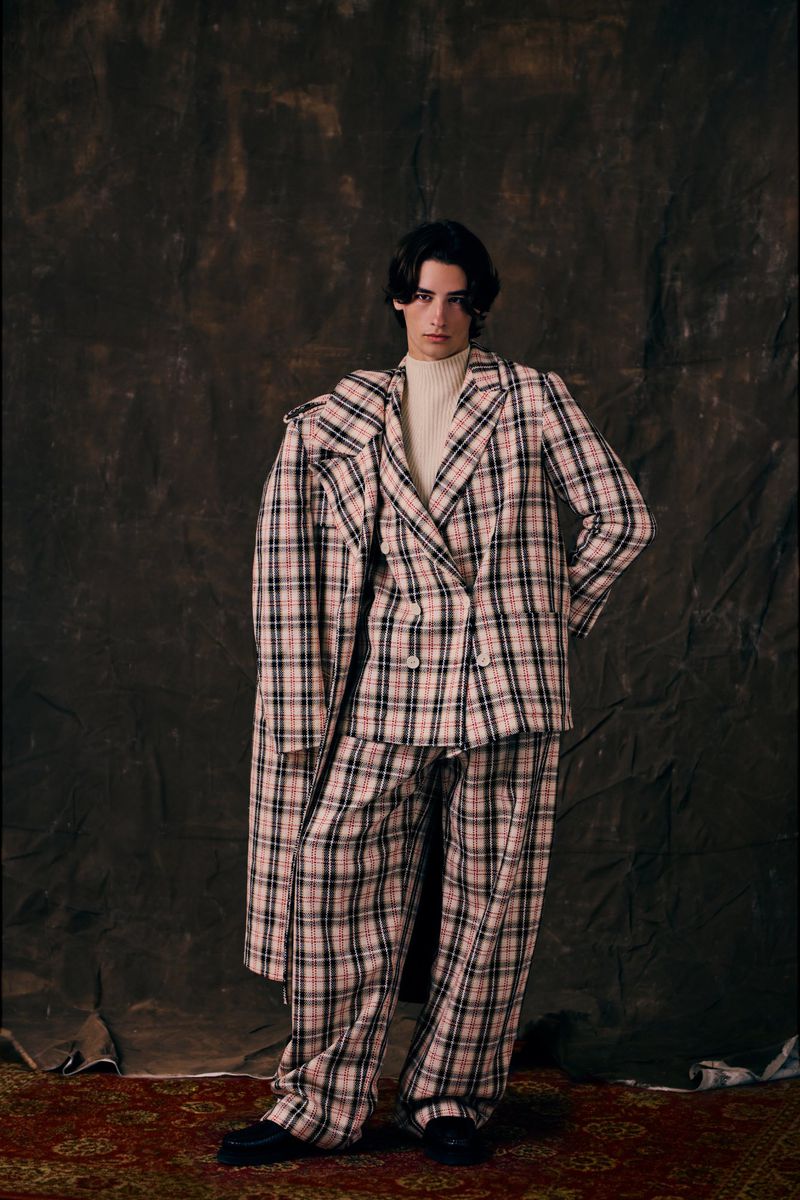 Male fashion model poses indoors wearing a tan and black plaid suit with an open coat, off-white turtleneck, and black dress shoes.
