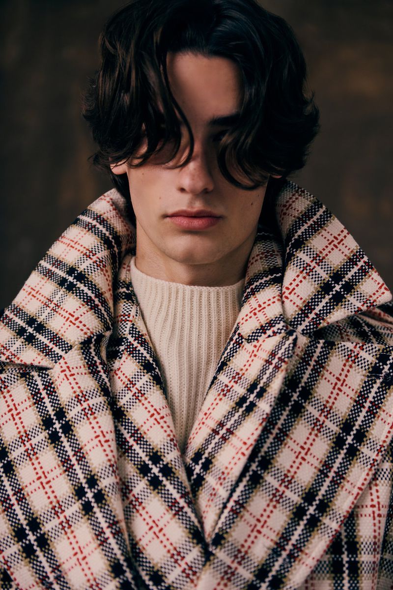 Portrait of a young adult male with dark hair, wearing a cream-colored sweater and a plaid wool coat, standing indoors against a dark backdrop.