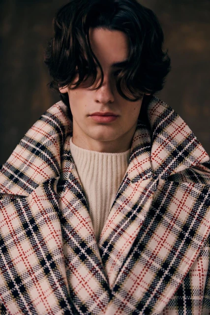 Portrait of a young adult male with dark hair, wearing a cream-colored sweater and a plaid wool coat, standing indoors against a dark backdrop.