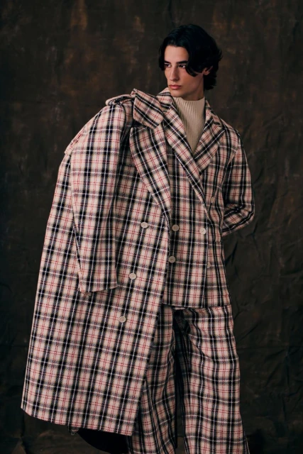 A person wearing a plaid pantsuit, indoors against a mottled backdrop. The outfit includes a long, double-breasted coat.