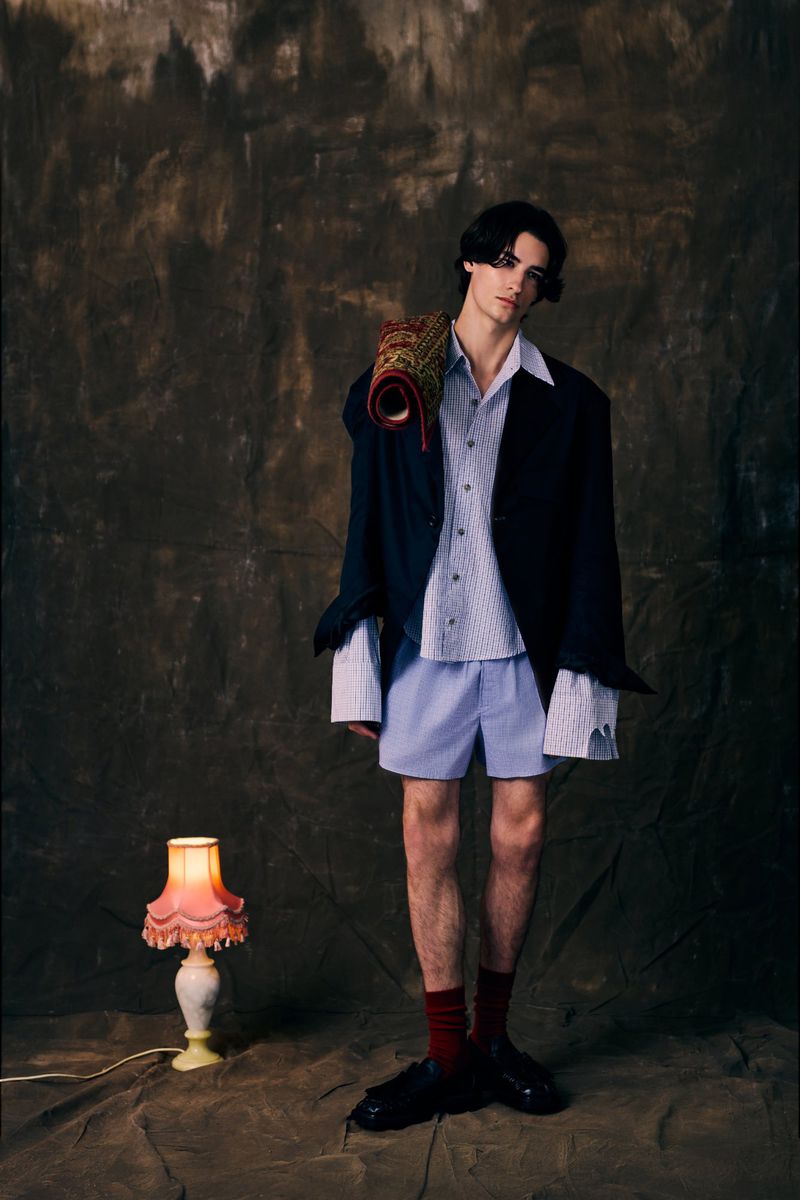 Full-length shot of a young adult male in a blue shirt and shorts, with a blazer and a rolled rug on his shoulder, indoors.