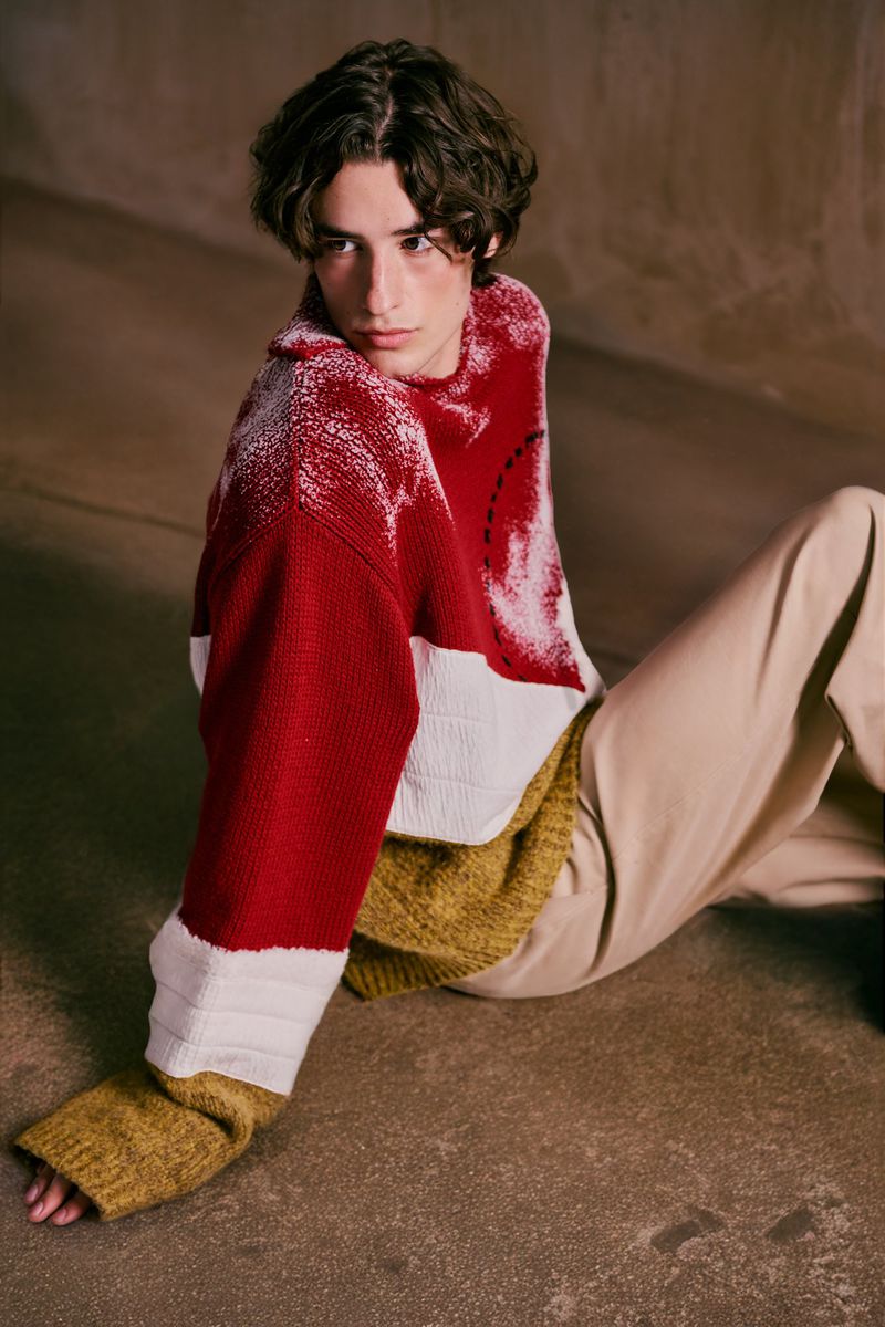 Young man seated on a concrete surface wearing a red, white, and mustard color-blocked knit sweater with beige trousers.