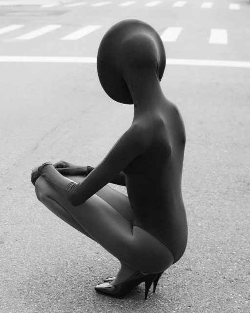 A crouched figure in a dark bodysuit and high heels is centered on an asphalt road, a monochrome outdoor fashion statement.