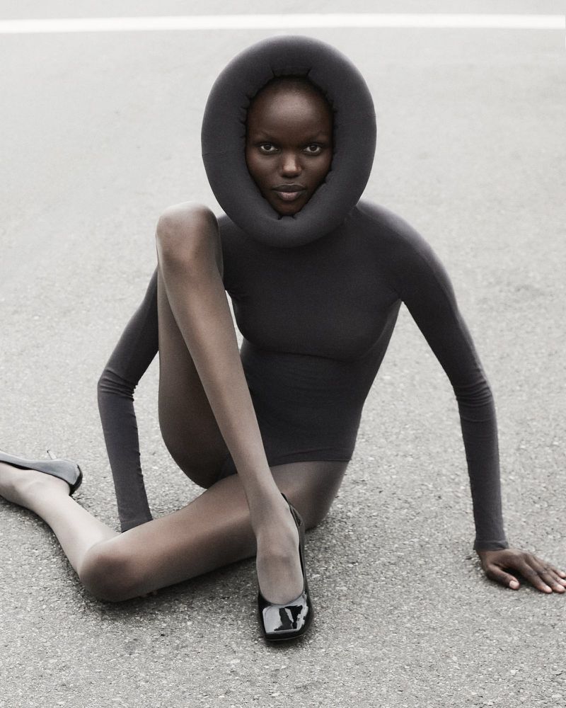 Fashion shot of a Black woman in a dark bodysuit with a padded neckpiece, sitting on asphalt wearing black patent flats.
