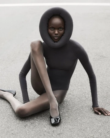 Fashion shot of a Black woman in a dark bodysuit with a padded neckpiece, sitting on asphalt wearing black patent flats.
