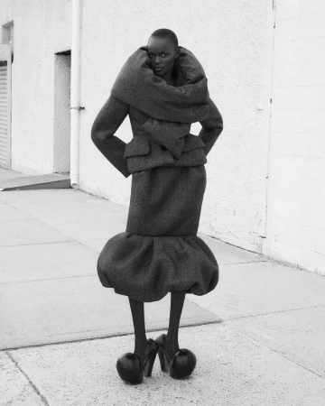 Striking black and white fashion shot of an African woman in an avant-garde jacket, skirt, and heels in an outdoor urban setting.