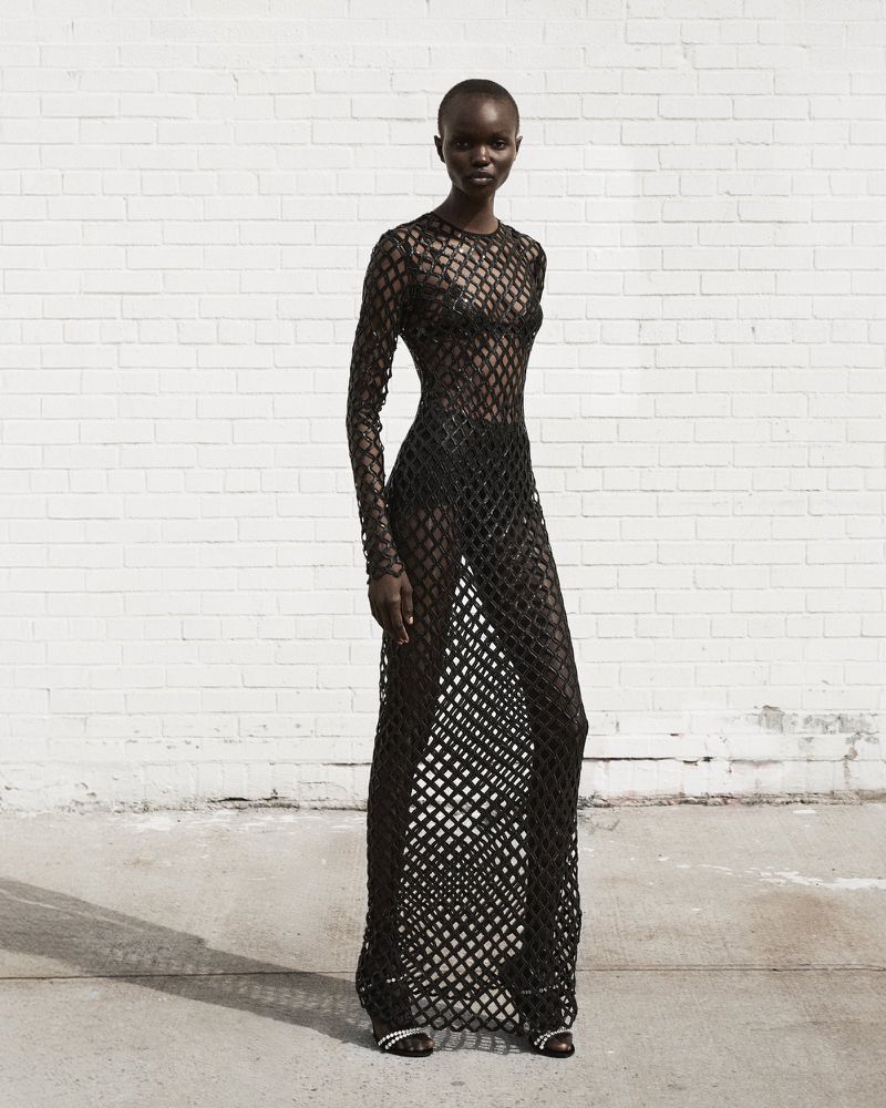 Fashion portrait of a black woman in a see-through black mesh dress, posing against a white brick wall with soft shadows, outdoors.