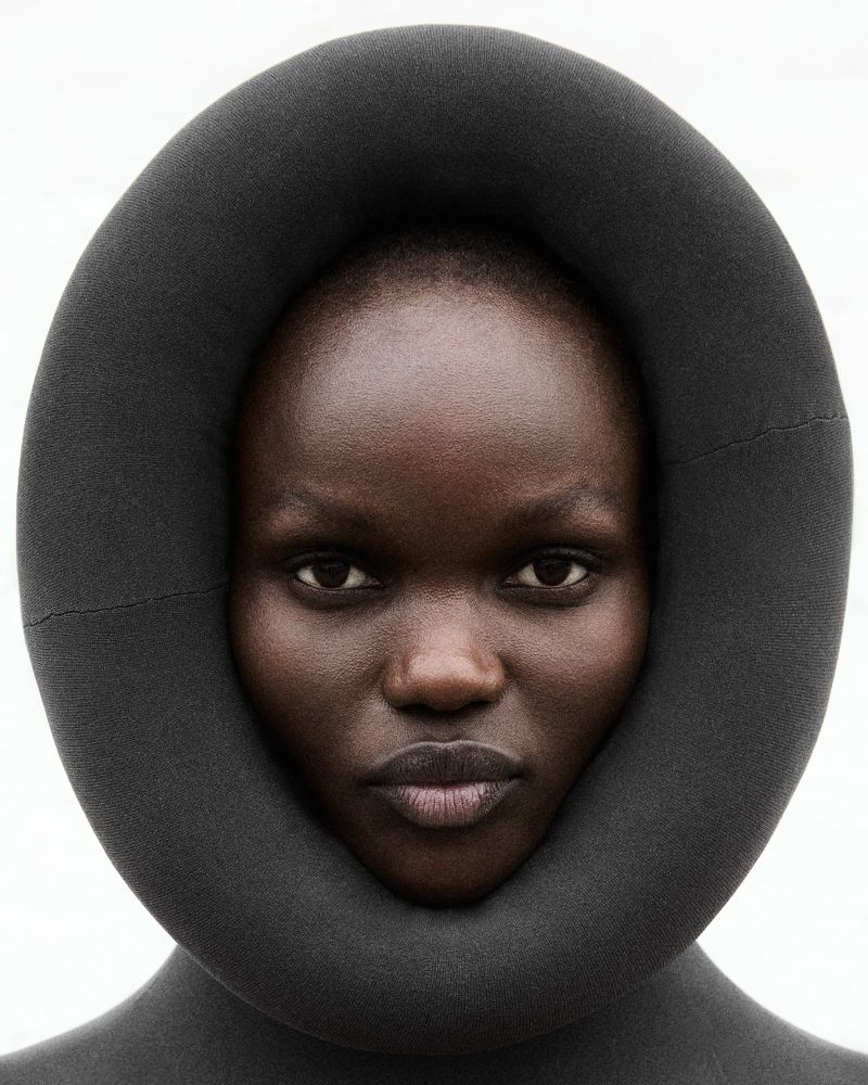 Close-up studio portrait of a Black woman wearing a voluminous, dark grey tubular garment framing her face. Minimal makeup, neutral expression.