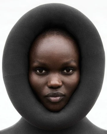 Close-up studio portrait of a Black woman wearing a voluminous, dark grey tubular garment framing her face. Minimal makeup, neutral expression.
