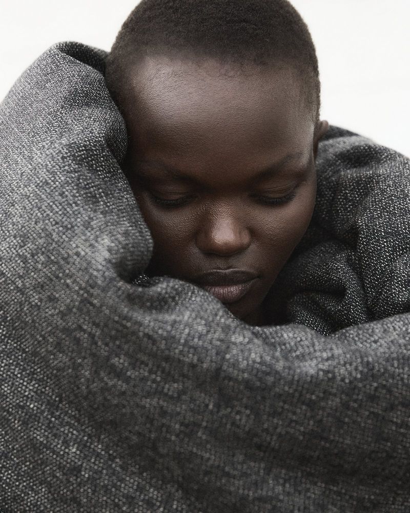 Close-up of an adult female with dark skin wrapped in a gray woven textile, eyes downcast, in a softly lit indoor setting.