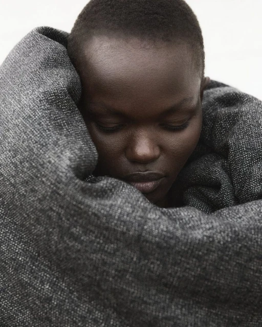 Close-up of an adult female with dark skin wrapped in a gray woven textile, eyes downcast, in a softly lit indoor setting.