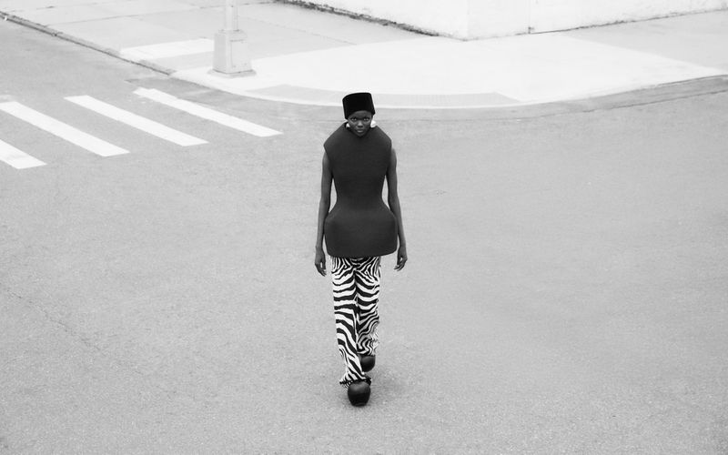 Woman walks on crosswalk wearing a black top, zebra print pants, a black hat, and oversized black platform shoes in monochrome.