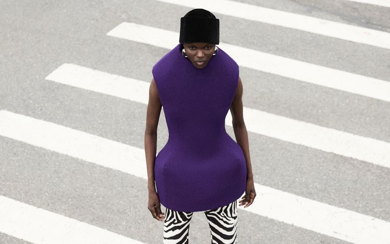 Fashionable woman with hat poses on pedestrian crossing wearing a purple sleeveless top and patterned black and white pants in street setting.