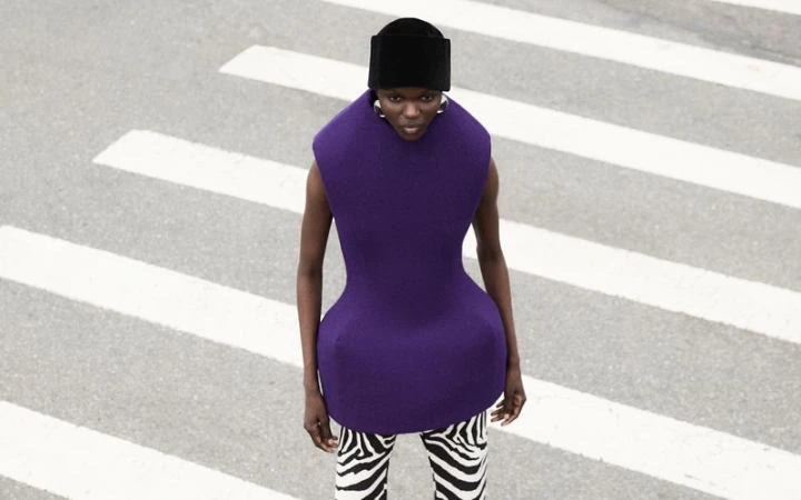 Fashionable woman with hat poses on pedestrian crossing wearing a purple sleeveless top and patterned black and white pants in street setting.