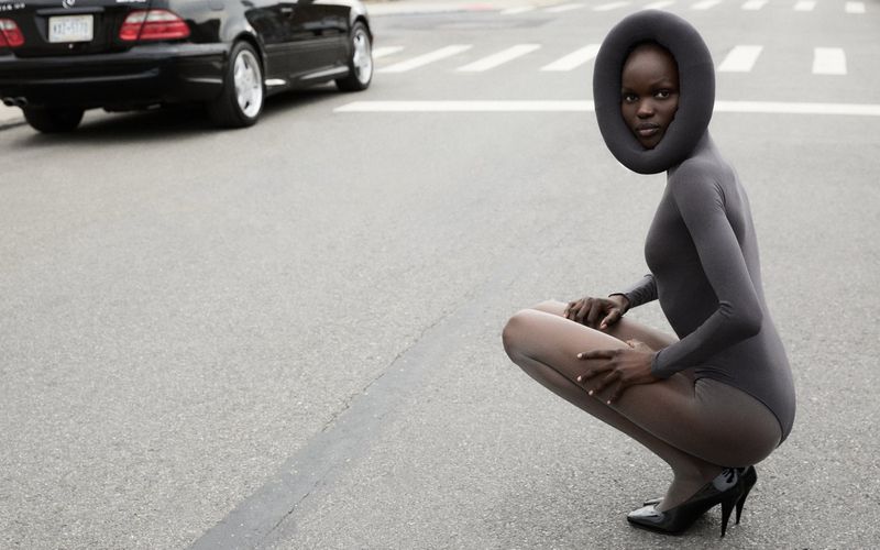 Adult female model in grey bodysuit and high heels poses on a grey road with a car in background wearing black padded headpiece.