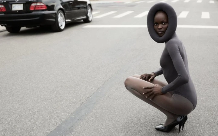 Model Adau Aguer crouching on an asphalt street in a grey bodysuit, sheer tights, black heels, and a padded circular headpiece.