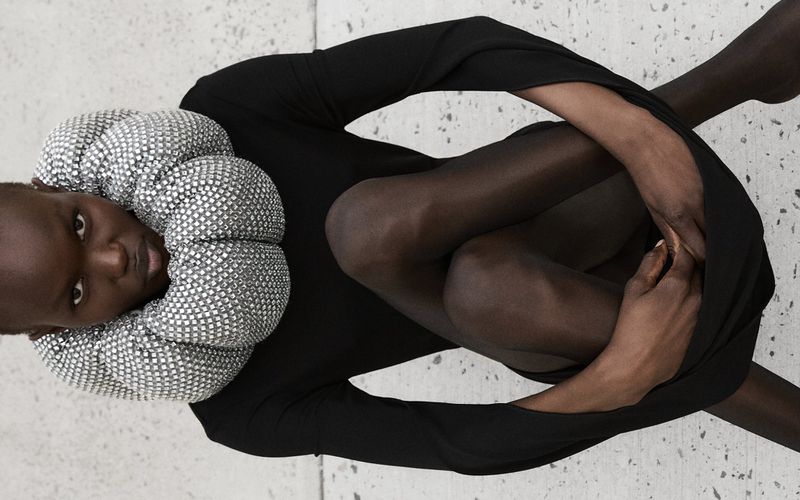 Fashion shot of a Black woman in a black long sleeve dress, posing while seated with legs and arms crossed, wearing a silver beaded collar.