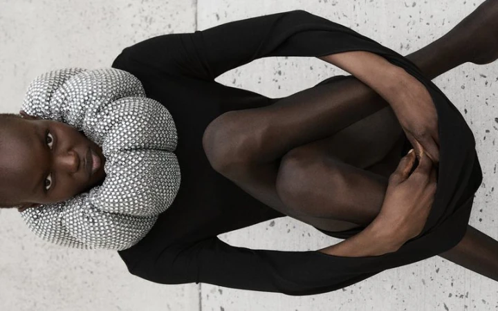 Fashion shot of a Black woman in a black long sleeve dress, posing while seated with legs and arms crossed, wearing a silver beaded collar.