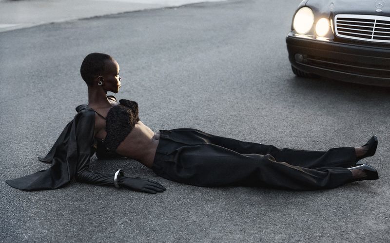 Woman in a black outfit poses on an asphalt road next to a parked black car with its headlights on, creating a striking, dramatic scene.