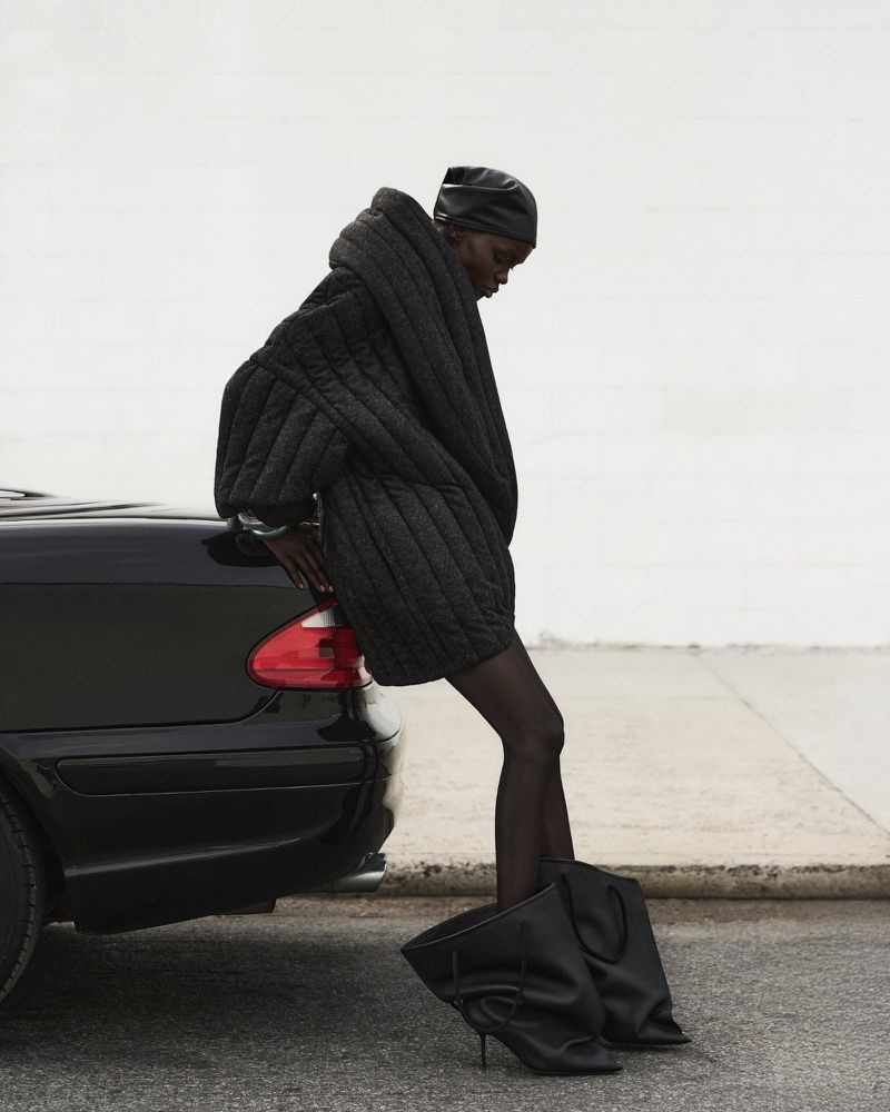 A slender woman wearing a black quilted jacket and matching headdress, leans on a black car parked on an outdoor city street.